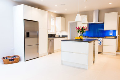 This is an example of a large contemporary u-shaped kitchen/diner in London with a double-bowl sink, flat-panel cabinets, white cabinets, composite countertops, blue splashback, glass sheet splashback, stainless steel appliances, porcelain flooring, an island, beige floors and grey worktops.