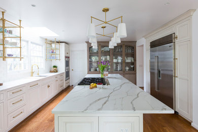 Photo of a large traditional u-shaped kitchen/diner in Cincinnati with a submerged sink, recessed-panel cabinets, white cabinets, quartz worktops, white splashback, ceramic splashback, stainless steel appliances, medium hardwood flooring, an island, brown floors and white worktops.