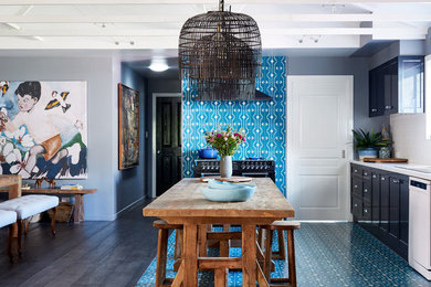 Cette image montre une cuisine américaine bohème en L avec un placard avec porte à panneau encastré, des portes de placard noires, une crédence blanche, un électroménager noir, îlot, un sol bleu et un plan de travail blanc.