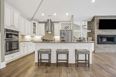 Open concept kitchen - mid-sized transitional l-shaped medium tone wood floor and brown floor open concept kitchen idea in Other with a farmhouse sink, granite countertops, beige backsplash, stone tile backsplash, stainless steel appliances, recessed-panel cabinets, white cabinets and an island