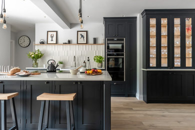Photo of a large classic u-shaped kitchen/diner in Surrey with a belfast sink, shaker cabinets, black cabinets, quartz worktops, white splashback, ceramic splashback, black appliances, ceramic flooring, an island and white worktops.