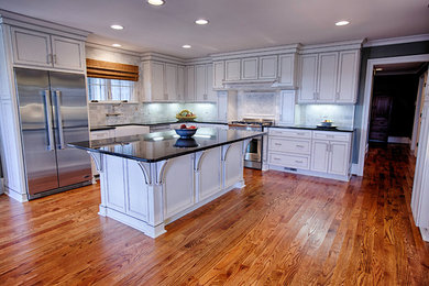 Inspiration for a mid-sized timeless l-shaped medium tone wood floor eat-in kitchen remodel in Birmingham with a farmhouse sink, flat-panel cabinets, white cabinets, granite countertops, gray backsplash, stone tile backsplash, stainless steel appliances and an island