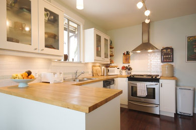 Eat-in kitchen - craftsman u-shaped dark wood floor and brown floor eat-in kitchen idea in Vancouver with a double-bowl sink, glass-front cabinets, white cabinets, wood countertops, white backsplash, subway tile backsplash, stainless steel appliances and a peninsula
