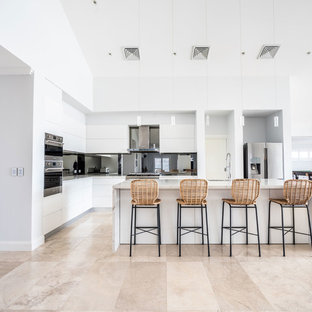 This is an example of a mid-sized contemporary l-shaped open plan kitchen in Sydney with flat-panel cabinets, white cabinets, mirror splashback, white benchtop, an undermount sink, quartz benchtops, metallic splashback, stainless steel appliances, with island and beige floor.