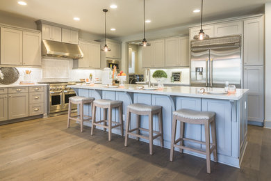 Example of a transitional l-shaped medium tone wood floor and brown floor kitchen design in Los Angeles with a farmhouse sink, shaker cabinets, gray cabinets, white backsplash, stainless steel appliances, an island and white countertops
