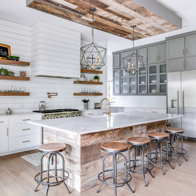 Example of a country l-shaped light wood floor and beige floor kitchen design in Dallas with a farmhouse sink, shaker cabinets, quartz countertops, white backsplash, wood backsplash, stainless steel appliances, an island, white countertops and white cabinets