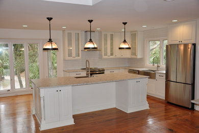 Example of a trendy medium tone wood floor kitchen design with a farmhouse sink, shaker cabinets, quartz countertops, white backsplash, subway tile backsplash, stainless steel appliances and an island