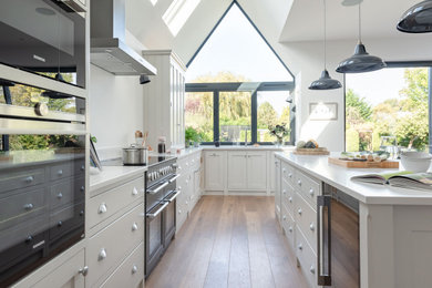 Photo of a large traditional u-shaped open plan kitchen in Surrey with shaker cabinets, white cabinets, quartz worktops, black appliances, light hardwood flooring, an island, brown floors and white worktops.