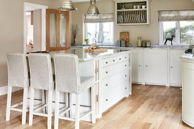 Design ideas for a large traditional l-shaped kitchen/diner in Wiltshire with a belfast sink, shaker cabinets, white cabinets, grey splashback, glass sheet splashback, white appliances, medium hardwood flooring and an island.