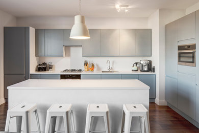 Design ideas for a contemporary l-shaped open plan kitchen in London with an integrated sink, flat-panel cabinets, blue cabinets, engineered stone countertops, white splashback, dark hardwood flooring, an island, stainless steel appliances and brown floors.