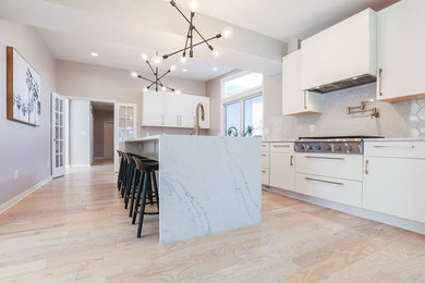 This is an example of a large contemporary l-shaped enclosed kitchen in Baltimore with a submerged sink, flat-panel cabinets, white cabinets, marble worktops, grey splashback, ceramic splashback, stainless steel appliances, light hardwood flooring, an island, beige floors and white worktops.