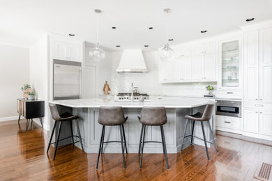 Example of a transitional l-shaped medium tone wood floor and brown floor open concept kitchen design in Vancouver with white cabinets, quartz countertops, white backsplash, subway tile backsplash, stainless steel appliances, an island, white countertops, an undermount sink and shaker cabinets