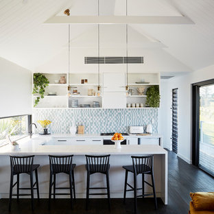 Beach style u-shaped open plan kitchen in Other with a farmhouse sink, flat-panel cabinets, white cabinets, multi-coloured splashback, panelled appliances, dark hardwood floors, a peninsula, black floor and white benchtop.