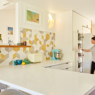 This is an example of an eclectic l-shaped eat-in kitchen in Los Angeles with a single-bowl sink, flat-panel cabinets, white cabinets, quartz benchtops, yellow splashback, ceramic splashback, stainless steel appliances, light hardwood floors, a peninsula, brown floor and white benchtop.