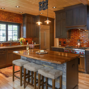 Foto de cocina en L, rural, de tamaño medio, abierta, con fregadero bajoencimera, armarios estilo shaker, puertas de armario de madera en tonos medios, electrodomésticos de acero inoxidable, suelo de madera clara, una isla, encimera de granito, salpicadero marrón, salpicadero de ladrillos y suelo beige