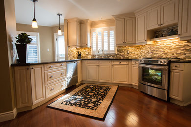 Mid-sized transitional l-shaped medium tone wood floor kitchen pantry photo in Toronto with an undermount sink, raised-panel cabinets, white cabinets, granite countertops, brown backsplash, glass tile backsplash, stainless steel appliances and a peninsula