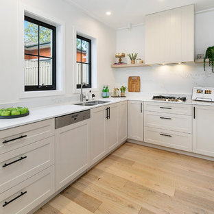This is an example of a small transitional l-shaped open plan kitchen in Sydney with a double-bowl sink, white cabinets, marble benchtops, marble splashback, stainless steel appliances, light hardwood floors, white benchtop, recessed-panel cabinets, white splashback, multiple islands and brown floor.