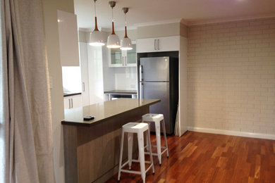 Kleine Moderne Wohnküche in U-Form mit flächenbündigen Schrankfronten, weißen Schränken, Granit-Arbeitsplatte, Küchenrückwand in Weiß, Küchengeräten aus Edelstahl, braunem Holzboden, Halbinsel und Rückwand aus Porzellanfliesen in Brisbane