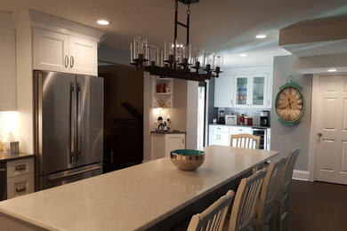 Example of a large transitional l-shaped dark wood floor and brown floor eat-in kitchen design in New York with a farmhouse sink, recessed-panel cabinets, white cabinets, quartz countertops, white backsplash, ceramic backsplash, stainless steel appliances and an island