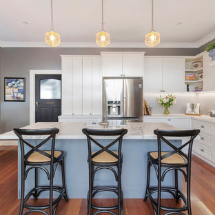 Mid-sized transitional l-shaped kitchen in Sydney with a farmhouse sink, shaker cabinets, white cabinets, quartz benchtops, grey splashback, ceramic splashback, black appliances, medium hardwood floors, with island, brown floor and white benchtop.