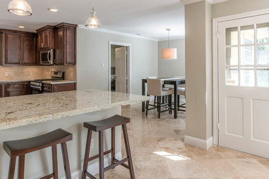 Example of a mid-sized classic u-shaped travertine floor eat-in kitchen design in Houston with flat-panel cabinets, dark wood cabinets, granite countertops, beige backsplash, stone tile backsplash, stainless steel appliances and an island
