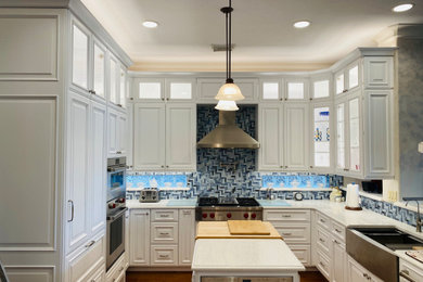 Large elegant u-shaped medium tone wood floor and brown floor open concept kitchen photo in Houston with a farmhouse sink, raised-panel cabinets, white cabinets, quartz countertops, blue backsplash, mosaic tile backsplash, stainless steel appliances, an island and white countertops
