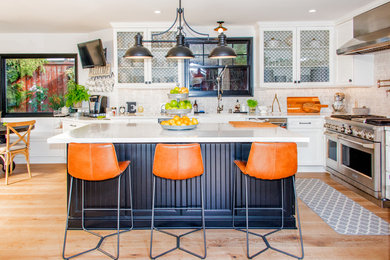 Example of a large transitional l-shaped medium tone wood floor and brown floor eat-in kitchen design in Los Angeles with a farmhouse sink, shaker cabinets, white cabinets, quartz countertops, white backsplash, porcelain backsplash, stainless steel appliances, an island and white countertops