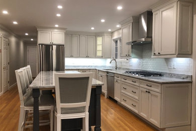 Example of a large transitional l-shaped medium tone wood floor and brown floor open concept kitchen design in Boston with an undermount sink, recessed-panel cabinets, white cabinets, quartz countertops, white backsplash, subway tile backsplash, stainless steel appliances, an island and multicolored countertops