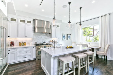 Example of a large transitional l-shaped dark wood floor and brown floor eat-in kitchen design in Orange County with a farmhouse sink, shaker cabinets, white cabinets, marble countertops, white backsplash, marble backsplash, stainless steel appliances and an island