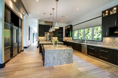 Design ideas for an expansive traditional l-shaped enclosed kitchen in Miami with flat-panel cabinets, dark wood cabinets, marble worktops, stainless steel appliances, multiple islands, a submerged sink, grey splashback, stone slab splashback and light hardwood flooring.