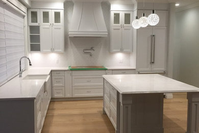 Inspiration for a large classic u-shaped kitchen in Vancouver with a belfast sink, recessed-panel cabinets, white cabinets, composite countertops, white splashback, integrated appliances, medium hardwood flooring, an island and brown floors.