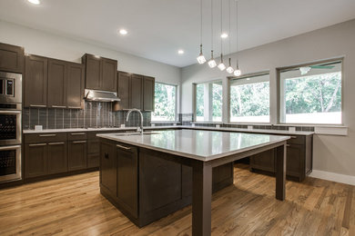 Example of a large minimalist l-shaped light wood floor open concept kitchen design in Dallas with a single-bowl sink, shaker cabinets, dark wood cabinets, granite countertops, brown backsplash, glass tile backsplash, stainless steel appliances and an island