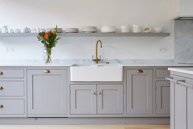 Inspiration for a medium sized contemporary l-shaped kitchen/diner in London with shaker cabinets, grey cabinets, composite countertops, white splashback, black appliances, an island, a belfast sink and medium hardwood flooring.