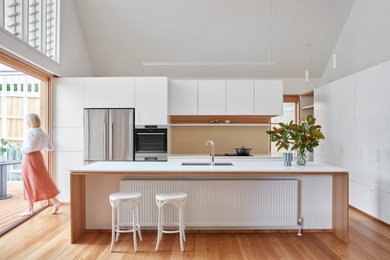 Example of a large trendy l-shaped medium tone wood floor, brown floor and vaulted ceiling eat-in kitchen design in Melbourne with a double-bowl sink, flat-panel cabinets, white cabinets, quartz countertops, stainless steel appliances, an island, white countertops and beige backsplash