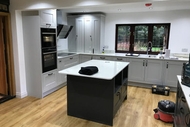 This is an example of a large classic kitchen/diner in Hertfordshire with a submerged sink, shaker cabinets, grey cabinets, quartz worktops, white splashback, glass sheet splashback, black appliances, vinyl flooring, an island, brown floors and white worktops.