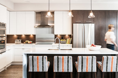 Modern inredning av ett kök, med en undermonterad diskho, släta luckor, vita skåp, brunt stänkskydd, rostfria vitvaror, mörkt trägolv och en köksö