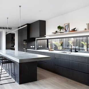 Photo of a modern galley kitchen in Melbourne with a double-bowl sink, concrete benchtops, light hardwood floors, with island, flat-panel cabinets, black cabinets, window splashback, black appliances, beige floor and grey benchtop.