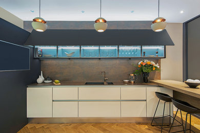 Example of a mid-sized trendy single-wall medium tone wood floor eat-in kitchen design in London with a drop-in sink, flat-panel cabinets, white cabinets, solid surface countertops, metallic backsplash, paneled appliances and a peninsula