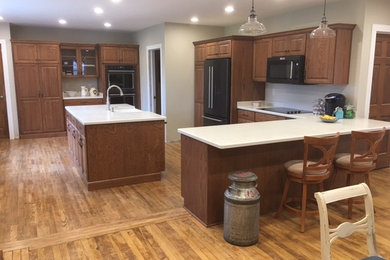 Example of a large classic u-shaped light wood floor eat-in kitchen design in Milwaukee with a farmhouse sink, raised-panel cabinets, medium tone wood cabinets, solid surface countertops, black appliances and an island