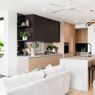 This is an example of a mid-sized contemporary l-shaped kitchen in Brisbane with an undermount sink, flat-panel cabinets, light wood cabinets, mirror splashback, stainless steel appliances, light hardwood floors, with island, beige floor and white benchtop.