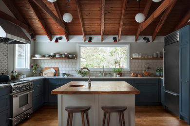 Example of a transitional u-shaped medium tone wood floor and brown floor kitchen design in Other with an integrated sink, shaker cabinets, blue cabinets, wood countertops, white backsplash, stainless steel appliances, an island and brown countertops