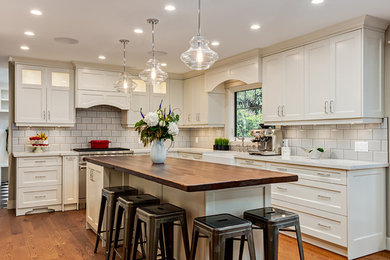 Example of a mid-sized transitional l-shaped light wood floor and beige floor enclosed kitchen design in Calgary with a farmhouse sink, recessed-panel cabinets, white cabinets, quartz countertops, white backsplash, porcelain backsplash, stainless steel appliances and an island