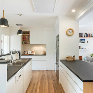 Design ideas for a mid-sized contemporary u-shaped open plan kitchen in Canberra - Queanbeyan with a double-bowl sink, flat-panel cabinets, white cabinets, beige splashback, ceramic splashback, stainless steel appliances, black benchtop, medium hardwood floors, a peninsula and brown floor.
