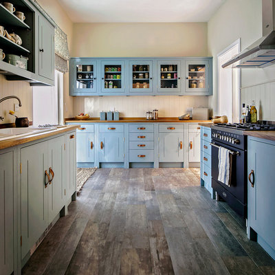 Example of a classic galley medium tone wood floor and brown floor enclosed kitchen design in London with a drop-in sink, shaker cabinets, blue cabinets, wood countertops, beige backsplash, black appliances and no island