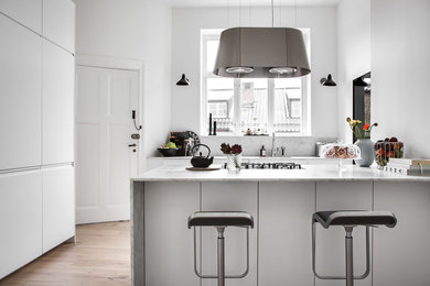 This is an example of a large scandinavian enclosed kitchen in London with an integrated sink, flat-panel cabinets, white cabinets, marble worktops, white splashback, marble splashback, black appliances, light hardwood flooring, an island and white worktops.