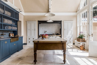 ロンドンにある高級な広いトラディショナルスタイルのおしゃれなキッチン (エプロンフロントシンク、落し込みパネル扉のキャビネット、青いキャビネット、白いキッチンパネル、磁器タイルのキッチンパネル、パネルと同色の調理設備、ライムストーンの床、ベージュの床、木材カウンター、茶色いキッチンカウンター) の写真