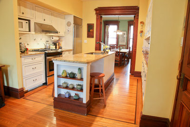 Example of a mid-sized ornate medium tone wood floor eat-in kitchen design in New York with an undermount sink, shaker cabinets, white cabinets, granite countertops, white backsplash, ceramic backsplash, stainless steel appliances and an island
