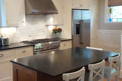 Example of a mid-sized transitional l-shaped medium tone wood floor enclosed kitchen design in Boston with an undermount sink, recessed-panel cabinets, white cabinets, soapstone countertops, white backsplash, stone tile backsplash, stainless steel appliances and an island