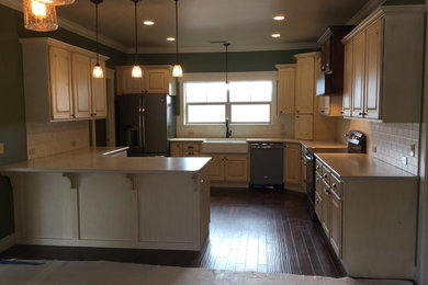 Example of a classic u-shaped dark wood floor enclosed kitchen design in Other with a farmhouse sink, raised-panel cabinets, distressed cabinets, solid surface countertops, white backsplash, subway tile backsplash, stainless steel appliances and a peninsula