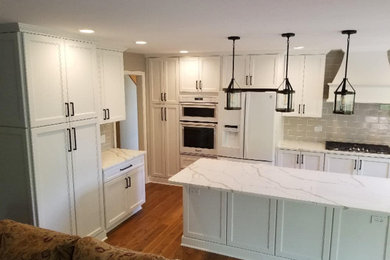 This is an example of a traditional kitchen in Chicago with a belfast sink, shaker cabinets, white cabinets, engineered stone countertops, grey splashback, metro tiled splashback, white appliances, medium hardwood flooring, an island, brown floors and white worktops.
